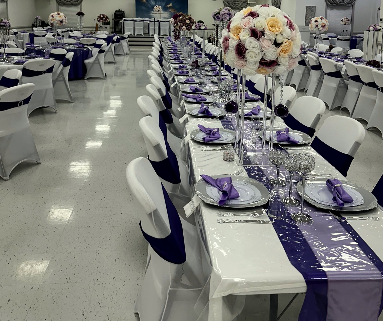 Formal purple, lavender and white reception environment with a long honored table, tall rose centerpieces and chair sashes, designed for a pastoral installation service with seating for more than 200 guests.